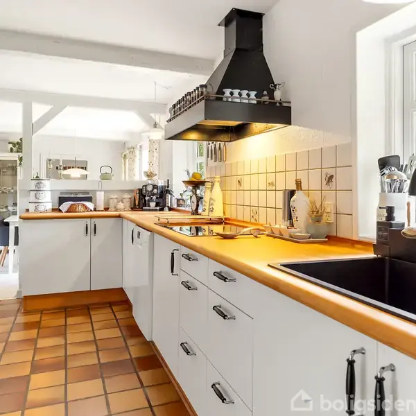 Et hvidt køkken med sorte hvidevarer og træbordplade, udsmykket med planter og køkkenredskaber, står i et lyst rum med brune fliser på gulvet og udsigt til haven gennem vinduet.