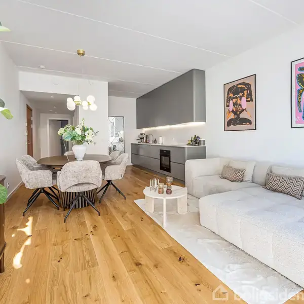 Et lyst og moderne opholdsrum med en stor hvid sofa, der står stille på et trægulv. Ved siden af er der et spisebord med blomster og vægkunst over sofaen.