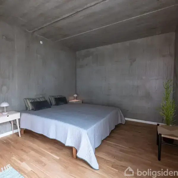 Seng dækket med gråt sengetæppe står i et minimalistisk soveværelse. Til venstre er der et natbord med lampe, til højre en plante og en stol, trægulv.