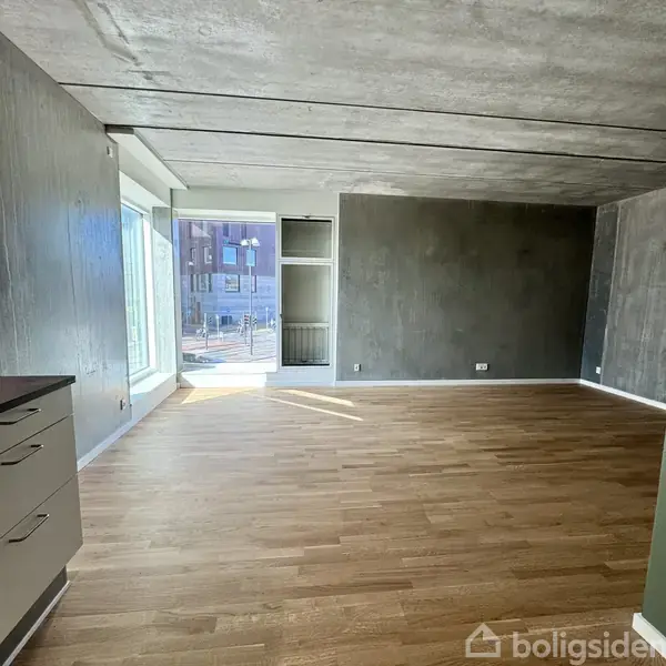 Et tomt rum med trægulv modtager lys fra store vinduer til venstre, viser en moderne bygning udenfor. Grå vægge og loft bidrager til en minimalistisk stil.