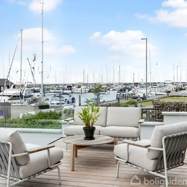 Terrassemøbler står på en træterrasse. De er arrangeret omkring et lavt bord med en plantepotte. Baggrunden viser en marina med fortøjede lystbåde under en blå himmel.