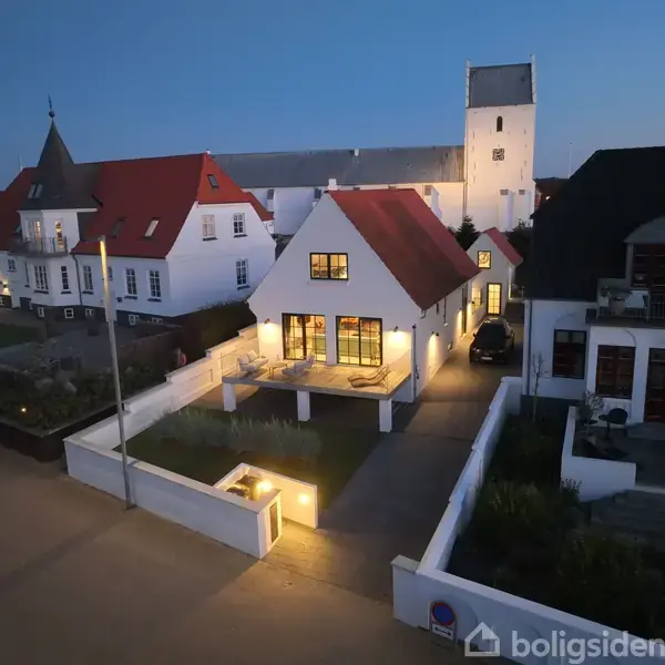 Et hvidt hus med rødt tag står oplyst om aftenen. Det har store vinduer med lys indefra. Omkring ses en have og naboliggende huse. En kirketårn er i baggrunden.