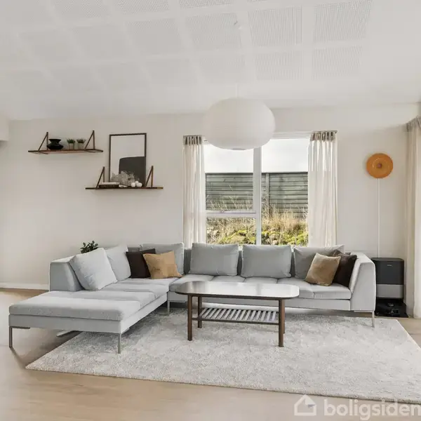 En grå sofa står i hjørnet af en stue på et lyst tæppe. Et sofabord står foran sofaen. Væggene har vinduer med gardiner og hylder med dekorationer.