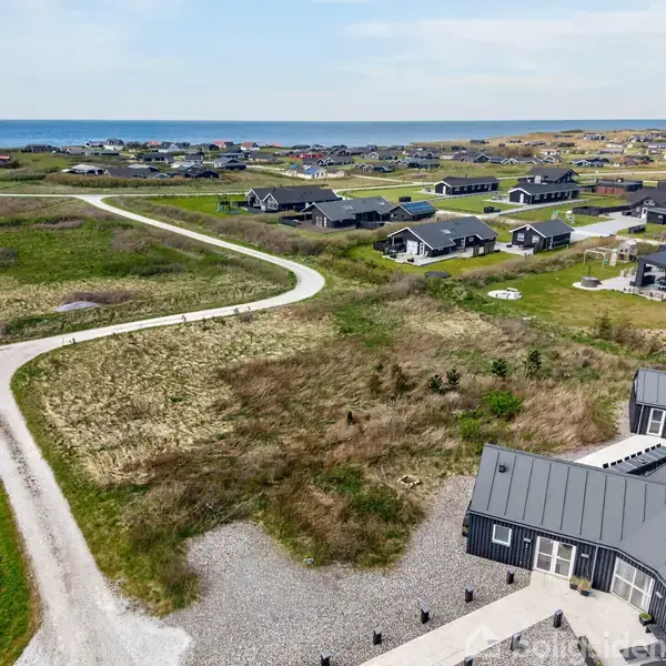 Flere sorte huse ligger spredt langs snoede stier i et grønt, åbent landskab tæt på kysten. Havet kan ses i baggrunden under en klar blå himmel.