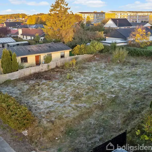 En lille bygning står stille ved siden af en åben, ujævn græsplæne i et boligområde med flere nærliggende huse og træer i efterårsfarver.