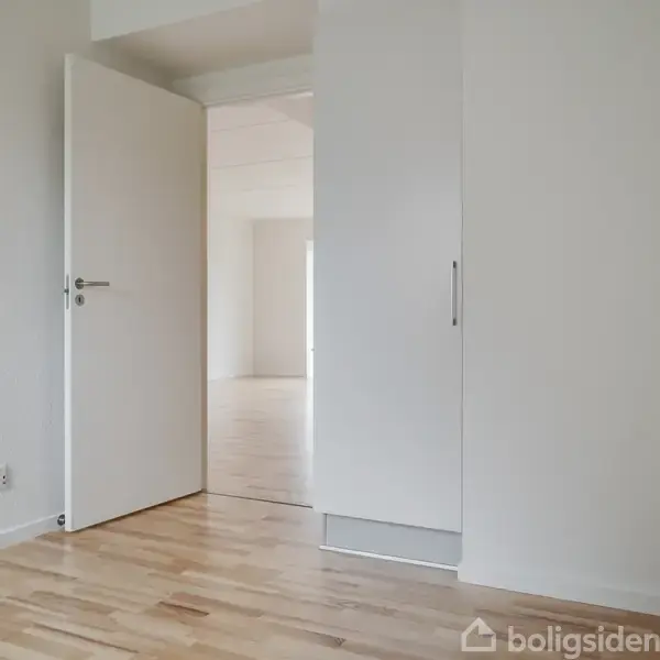 En åben dør leder ind til et lyst rum med lys trægulv og hvide vægge. Konteksten er et minimalistisk interiør uden møbler, hvilket skaber en rummelig atmosfære.