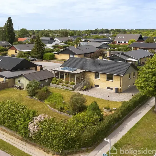 Et parcelhus med sort tag står i et stille boligkvarter. Huset har en velplejet have omgivet af hække og ligger på et hjørne ved en asfalteret vej.