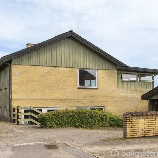 Opgang til gul murstensvilla med grøn beklædning, omgivet af lav hæk og garagebygning. Beliggende på en stille vej, fremhæver husets få vinduer et typisk skandinavisk design.