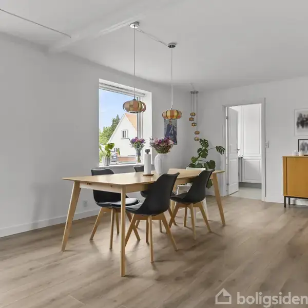 Spisebord med tre stole står midt i et lyst rum med trægulve. To vinduer giver naturligt lys. En kommode står nær en dør, dekoreret med planter og billeder.