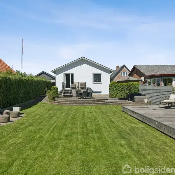 Et hvidt hus står i en velplejet have med grøn græsplæne. Foran huset er der en terrasse med havemøbler. Omkringliggende huse og hegn skaber en hyggelig boligkvarterstemning.