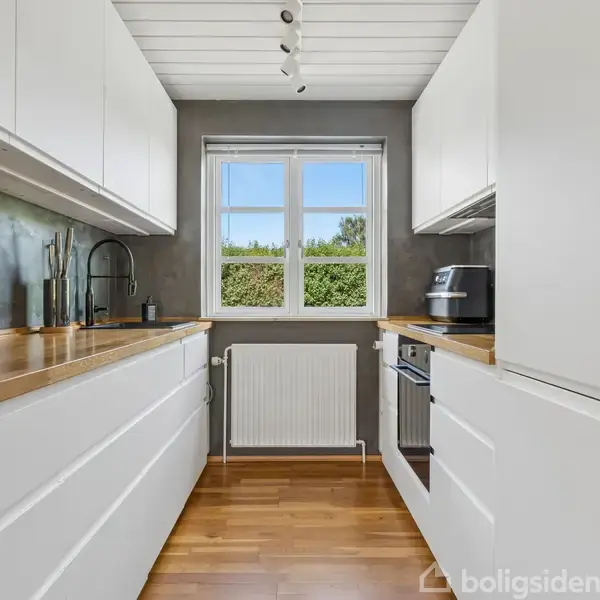 Køkken med hvide skabe strækker sig langs begge sider. En ovn og vask ses til højre, med et vindue i centrum. Trægulve og grøn udsigt udenfor.