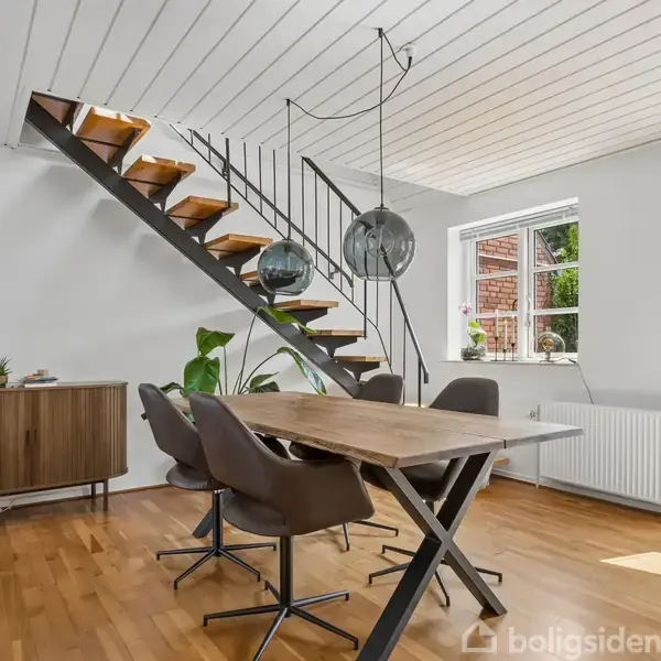 Spisebord med brune stole står under en moderne trappe med åbne trin. Rummet har hvidt loft, store vinduer, lys træparket og minimal dekoration med grønne planter og en skænk.