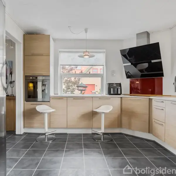 Et moderne køkken med lyse træskabe og en stor vægmonteret sort tv-skærm. To hvide barstole står ved køkkenbordet. Stor grå flisebelægning i et åbent rum.