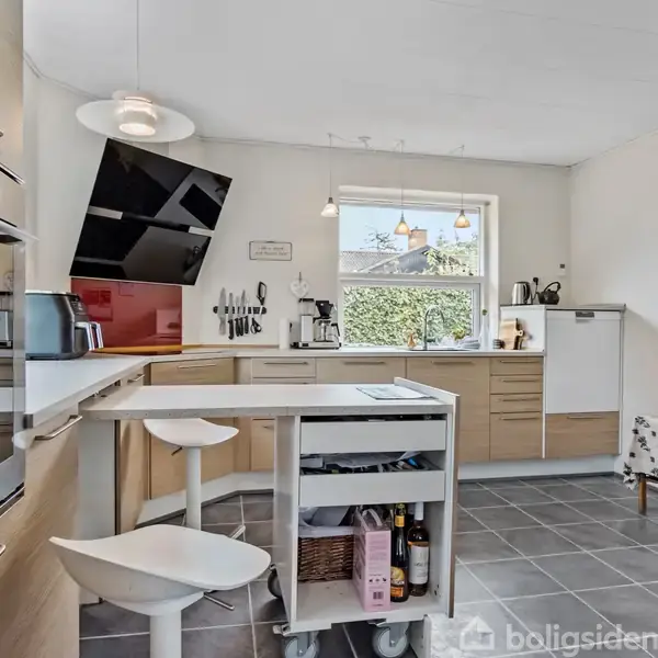 En moderne køkkenø med to barstole står i centrum. Køkkenet har træskabe, et stort vindue, samt diverse køkkenredskaber. Væguret viser klokken. Vindue ud mod en hæk.