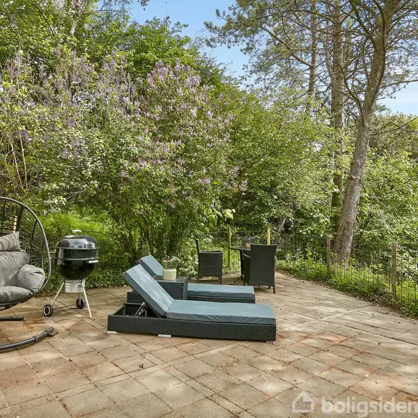 Havemøbler står på en solbeskinnet terrasse med en hængestol og grill. Omkring er der grønne træer og buske, der skaber en frodig og afslappet atmosfære.