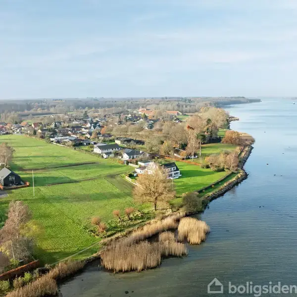 En samling huse grænser op til en stor sø eller fjord, omgivet af grønne marker og træer. Himlen er klar, og vandet ser roligt ud.