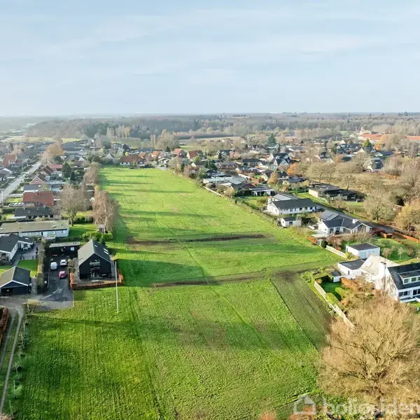 Grønt engområde strækker sig gennem en lille by med spredte huse. Omkringliggende træer og huse markerer byens grænser, mens veje forbinder forskellige kvarterer under en klar himmel.