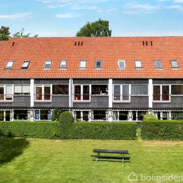 Rækkehus bygning med rødt tegltag og store vinduer langs facaden, omgivet af grønne hække og en græsplæne med en bænk.