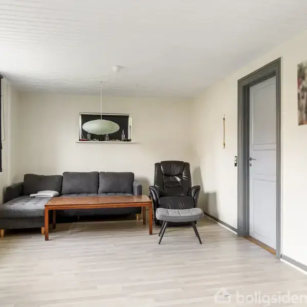 Grå sofa står mod en væg med et spejl over, mens en lænestol med skammel er placeret ved siden af. Rummet har lyse vægge og gulv samt et stort vindue til venstre.