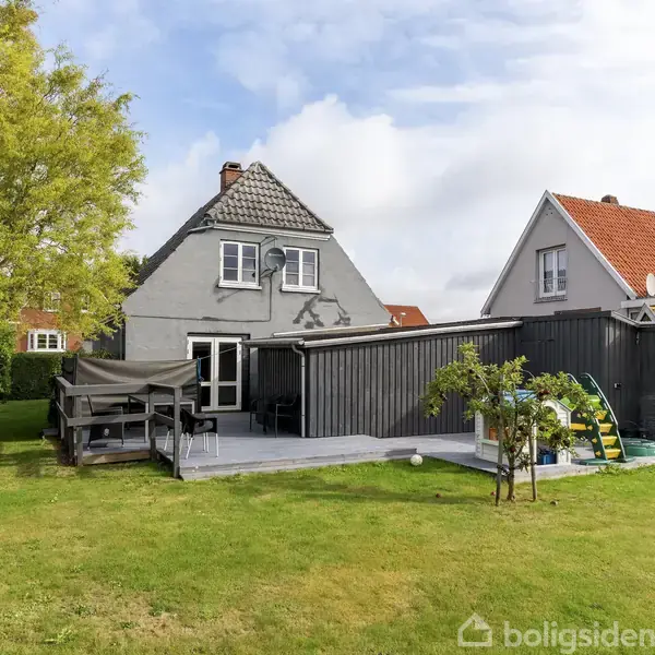 Et gråt hus med en terrasse ses fra haven; havemøbler er placeret på terrassen. Ved siden af bygges et lavt, sort skur. Græsplæne og et stort træ omkranser området.