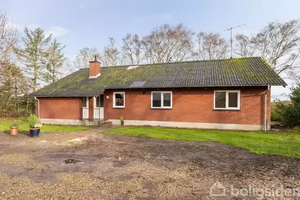 Et rødt murstenshus med mosgroet tag står i en åben, jordet have omkranset af træer. En stor krukke med planter står ved siden af den lille trappe ved hoveddøren.