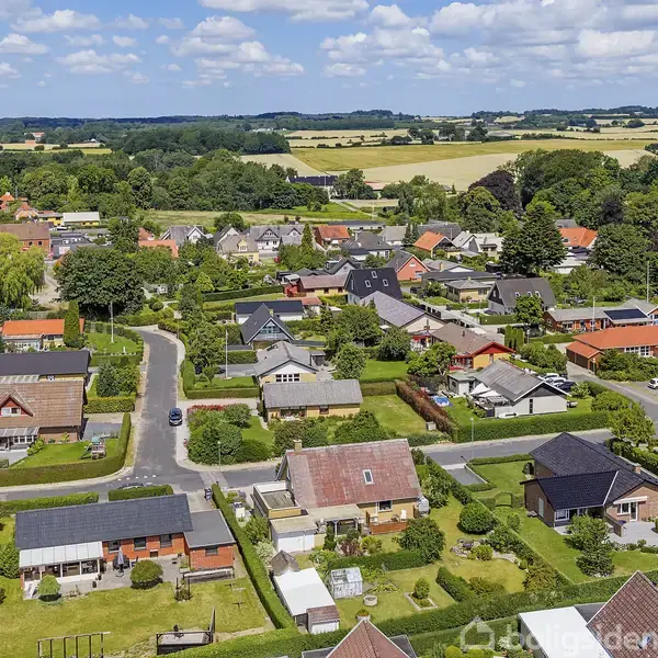 Bebyggelse bestående af enfamiliehuse med røde tage omgivet af velplejede haver og træer. Nabolaget omkranses af marker og skov, under en lysblå himmel med spredte skyer.