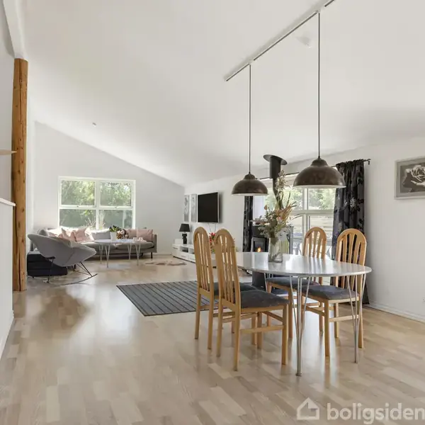 En lys stue med spisebord og træstole står midt i rummet. I baggrunden er der en sofa ved et stort vindue. Rummet har trægulve og moderne indretning.