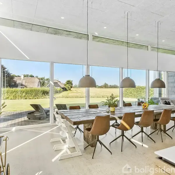 En stor spisebord med moderne stole står i centrum. Bordet er dekoreret med blomster. Over bordet hænger tre pendellamper. Rummet har store vinduer med udsigt til en have.