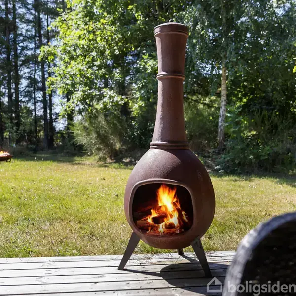 Chimenea med ild brænder på en terrasse, omgivet af græsplæne og træer i sollys.