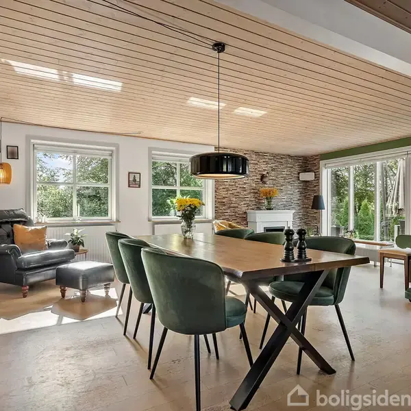 Et moderne spisebord med grønne stole står i en lys stue. Rummet har træloft, store vinduer og en pejs ved en stenbeklædt væg. Blomster pryder bordet.