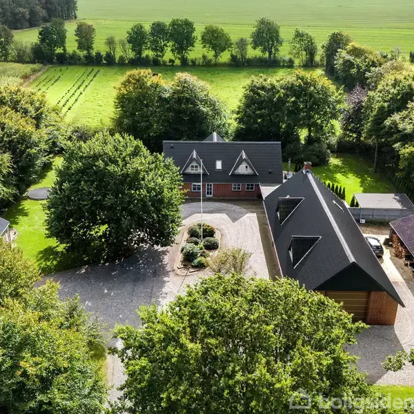 Stort hus med spidse tage set fra luften, omgivet af træer og grønne marker. Gårdspladsen er centralt placeret, med flere bygninger rundt om den.