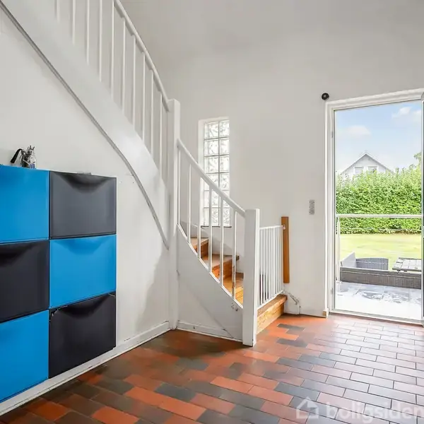 Trappe med hvidt gelænder leder op, mens blå sorte skabe hænger på væggen; dør åben til have belagt med mursten.