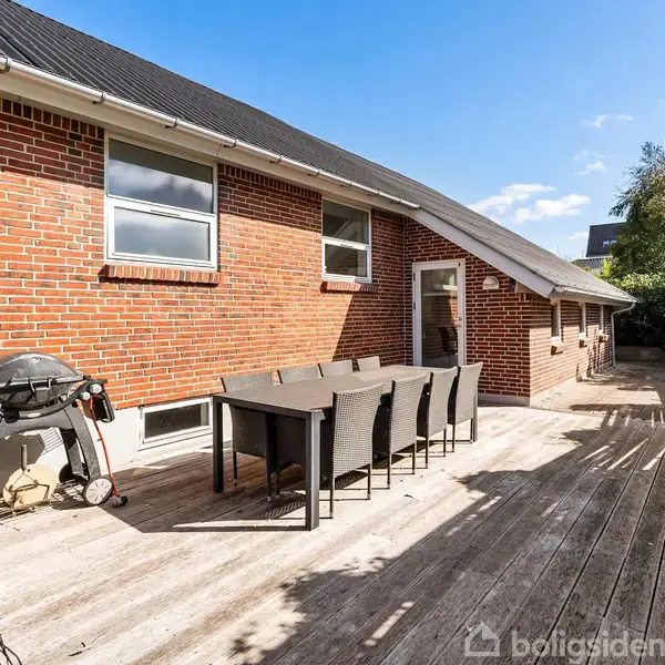 Grill og spisebord med stole står på en træterrasse foran en murstensbygning. Terrasse er delvist skygget, omgivende buske og en klar blå himmel.