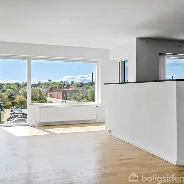 Et lyst rum med trægulv og store vinduer, der indrammer en udsigt over et grønt landskab. En halvmur adskiller rummet, og radiatorer er placeret under vinduet.