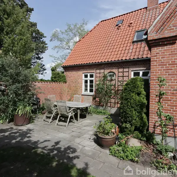 Et bord med stole står på en terrasse. Omkranset af potteplanter og tæt på et rødt murstenshus med rødt tegltag. Træer og buske i baggrunden.