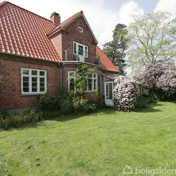 Et rødt murstenshus med rødt tegltag står i en frodig have. Huset har hvide vinduesrammer, en altan, og en rig vegetation omgiver det med buske og træer.