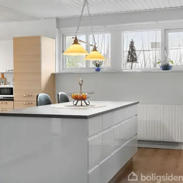 Et lyst køkken med en stor, hvid ø i midten, dekoreret med et frugtfad. Over øen hænger to gule lamper. Vinduer i baggrunden giver naturligt lys.