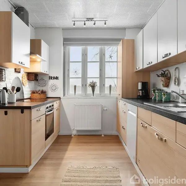 Et moderne køkken med træskabe og sorte bordplader. Køkkenapparater som ovn og mikroovn integreret. Et vindue ved enden med udsigt, dekorativ vægtekst "HOME" og planter på hylden.