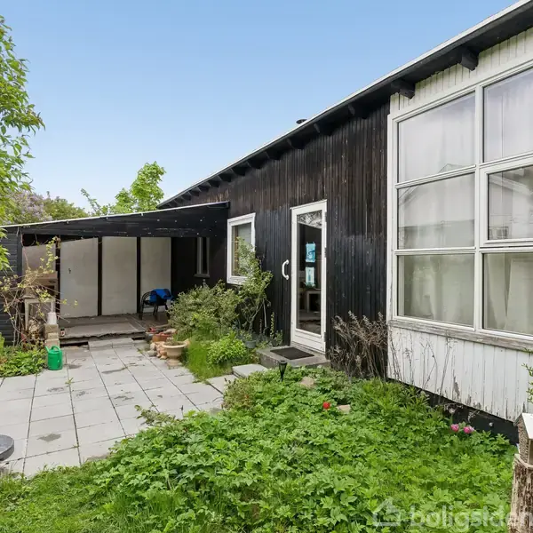 Sort træhus med store vinduer, der åbner ud mod en have med fliser og grønne buske. En overdækket terrasse er synlig i baggrunden med nogle redskaber og objekter på gulvet.