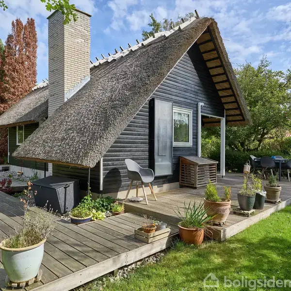 Stråtækt hus står på en træterrasse. Husets mørke facade er omringet af krukker med planter. Grønt græs og buske omgiver terrassen under en blå himmel med få skyer.