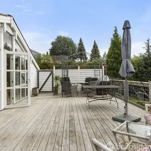 Træterrasse med havemøbler og en paraply står stille under åben himmel. Omgivet af grønne buske og træer, er terrassen tilknyttet et hus med store glasfacader.