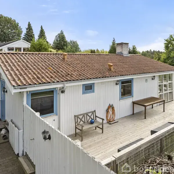 Et hvidt hus med rødbrunt tag stående på en træterrasse. Bord og bænk er placeret langs væggen. Haven er omgivet af træer, der skaber et grønt miljø.