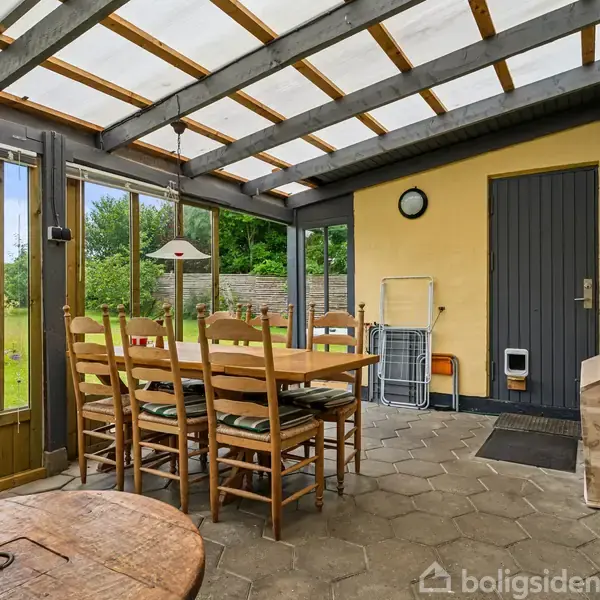 Et spisebord med stole står i en overdækket veranda med store vinduer. En grå dør leder udenfor, og i hjørnet er en trækasse og en vægur. Græsplæne udenfor.