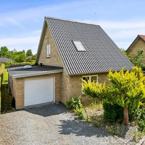 En murstenshus med sort tag står stille i et villakvarter. Grusvej fører til garagen. Omkranset af grønne buske og en frodig have.