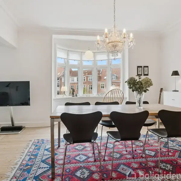 Et spisebord med sorte stole står under en lysekrone. En stor karnap vindue viser bygninger udenfor. Et tæppe er under bordet, og et fjernsyn står til venstre.