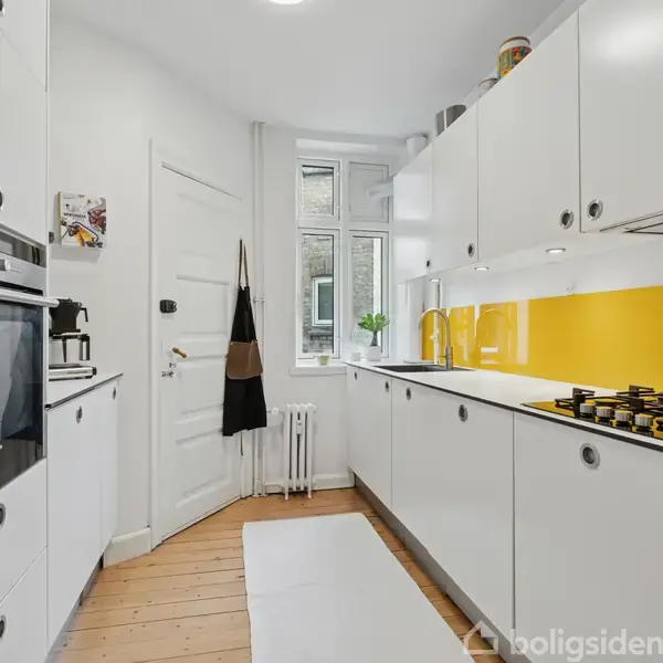 Et lyst, moderne køkken med hvide skabe, gul bagvæg og en gasblusplade. En ovn er indbygget på venstre side. Et vindue i baggrunden lader naturligt lys ind.