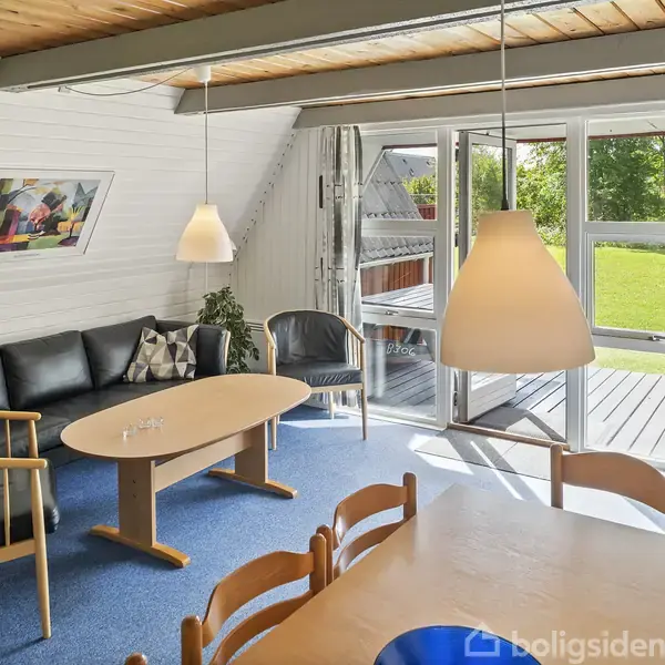 En stue med sort lædersofa og træbord står ved siden af vinduer med udsigt til en grøn have. En blå tæppe dækker gulvet, og hvide vægge omkranser rummet.