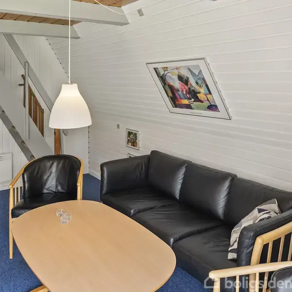 Sort lædersofa står i en stue med blåt gulvtæppe, en træsofabord og en sort lænestol. Et billede hænger på den hvidmalede væg, og en lampe hænger ned fra loftet.