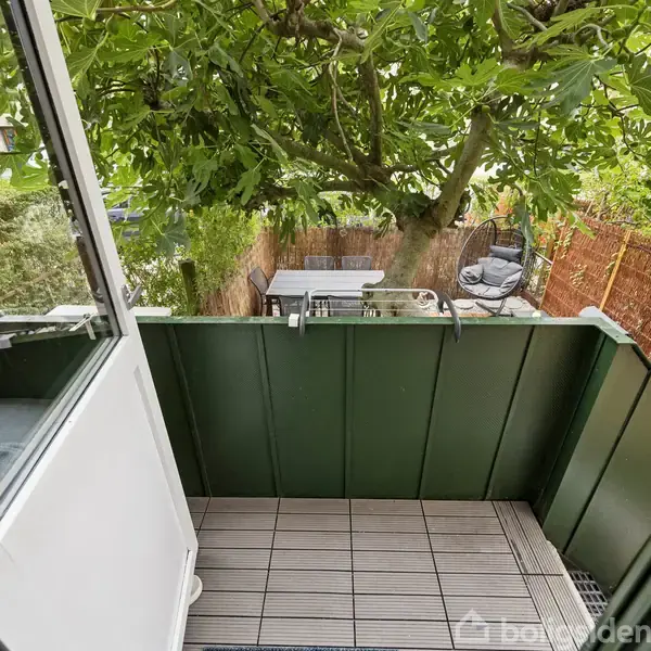 En lille balkon med trælaminatgulv åbner ud mod en frodig have med et stort træ. Et bord og stole ses i baggrunden.