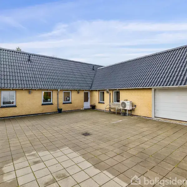 Et stort, gult murstenshus med sort tegltag står stille ved en stor brolagt gårdsplads. To vinduer og en hvid garageport ses ved siden af en udendørs varmepumpe.